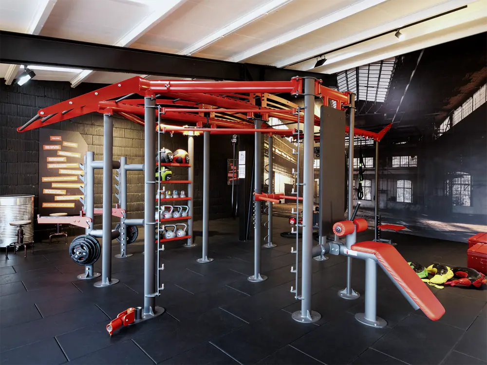 Rotes Functional-Rig mit Regal für Kettlebells und Medizinbälle, Hantelscheiben und Bauchbank vor einer Fototapete mit Industriehalle