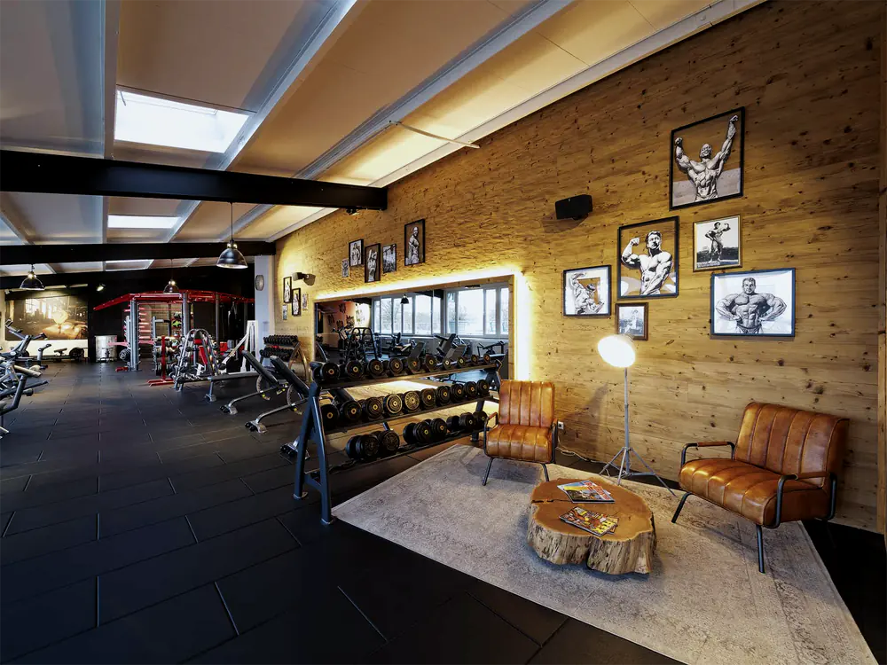 Kurzhantelregal vor der beleuchteten Spiegelwand, rechts eine Sitzecke mit Ledersesseln und gerahmten Bodybuilding-Zeichnungen an der Holzwand