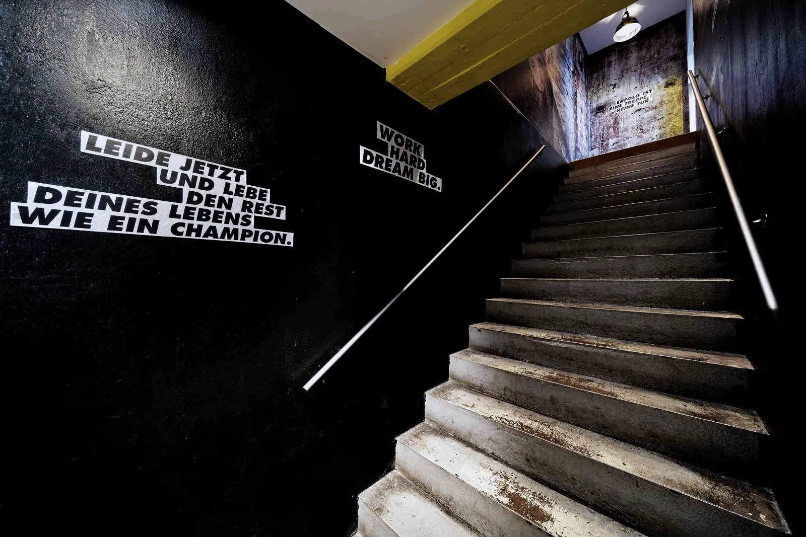Treppenaufgang im TC Training Center: schwarze Wand mit dem Schriftzug „Work hard, dream big.“, oben eine Industriehallen-Tapete